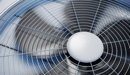 A close-up, high-angle shot shows the spinning blades of an air conditioning unit's condenser fan behind a circular metal safety grille.