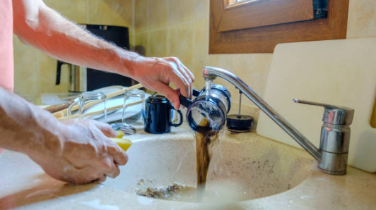 A person rinses a French press under a running faucet, pouring dark coffee grounds and liquid directly into a kitchen sink.