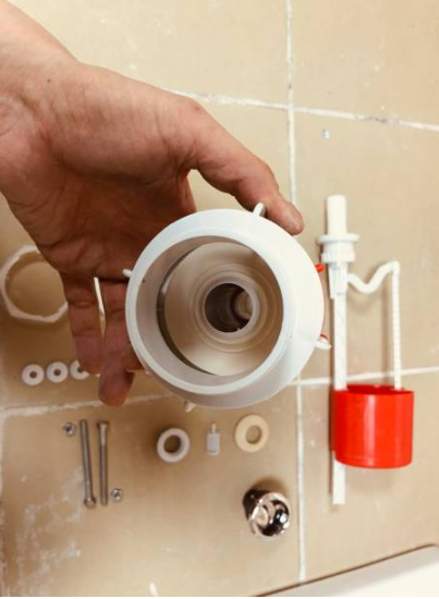 A hand holds a white plastic flush valve component over a tiled surface displaying various toilet repair parts, including a red float cup and mounting bolts.