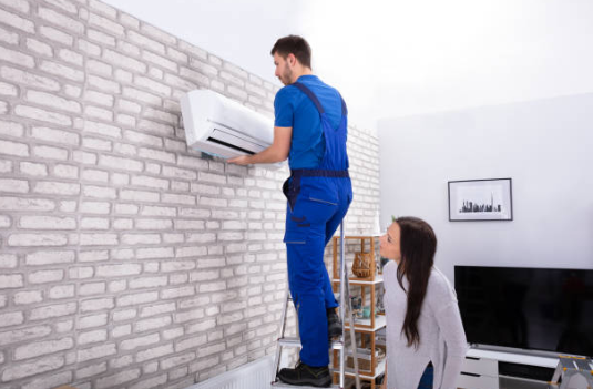 A technician in blue overalls stands on a ladder to install a white indoor mini-split air conditioning unit onto a light-colored brick wall while a homeowner watches.