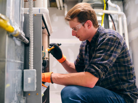 A technician wearing safety glasses and orange-accented gloves uses a small flashlight to inspect the internal components of a grey residential furnace.