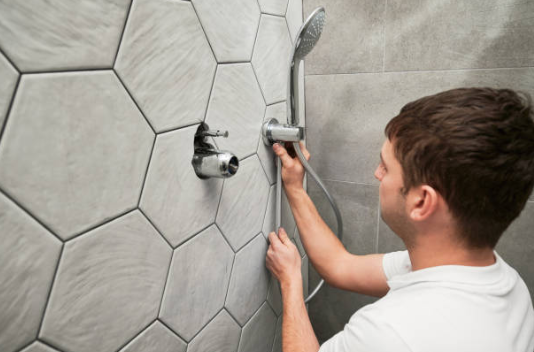 A man in a white t-shirt installs a chrome handheld shower head and hose onto a modern grey hexagonal tiled wall.