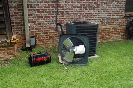 An outdoor air conditioning condenser unit sits disassembled on a lawn next to a brick wall, with its fan motor assembly removed and a red tool bag resting nearby.