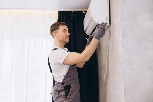 An HVAC technician wearing grey overalls and protective gloves carefully mounts a white indoor mini-split air conditioning unit onto a living room wall.