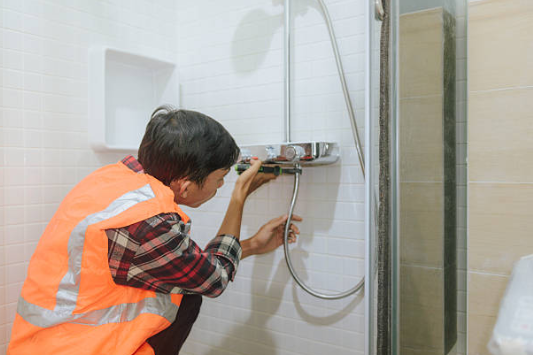 A technician wearing a high-visibility orange safety vest uses tools to install or repair a chrome shower faucet and hose against a white tiled wall.