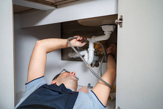 A plumber wearing glasses and blue overalls works inside a kitchen cabinet to connect flexible water supply lines to the pipes beneath a sink.