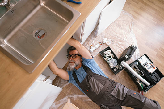 A plumber lying on the floor repairing pipes under a kitchen sink with tools and plumbing parts placed nearby.