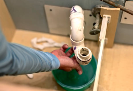 A plumber's hand reaches toward a disassembled white PVC P-trap under a sink, positioned over a green bucket catching residual water.