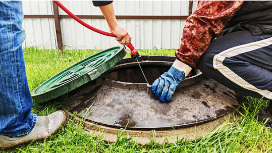 Workers opening a septic tank lid and using a hose to clean or inspect the system in an outdoor residential area.