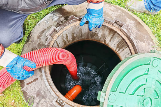 A worker using a large hose to pump out waste from an open septic tank during a cleaning or maintenance process.