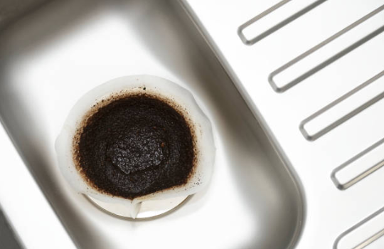 A top-down view shows dark coffee grounds collected in a white paper filter sitting inside a stainless steel kitchen sink drain.