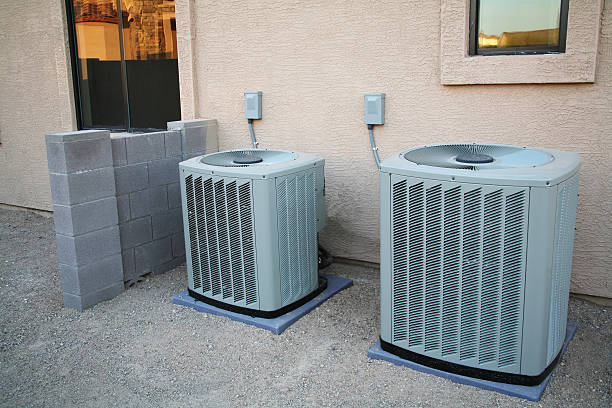 Two air conditioning units on pads beside a stucco wall with a cinder block screen