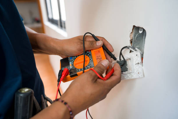 Electrician using an orange digital multimeter to test the wiring of an open electrical outlet on a wall