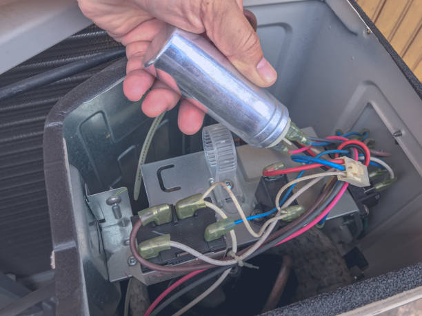 A technician holding a replacement HVAC capacitor while servicing the electrical components inside an outdoor air conditioning unit to restore proper system operation.