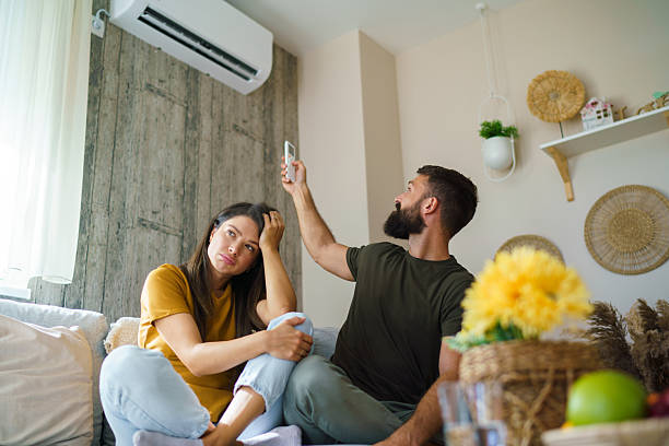 A homeowner using a remote control to adjust a wall-mounted air conditioner while another person appears frustrated, highlighting common home cooling and air conditioning performance issues.