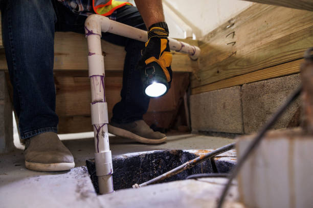 Plumber holding a flashlight while inspecting a sump pit with PVC discharge piping in a crawl space or basement.