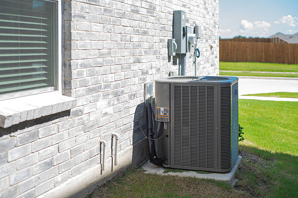 Outdoor air conditioner condenser unit against a gray brick house wall with an electrical disconnect box