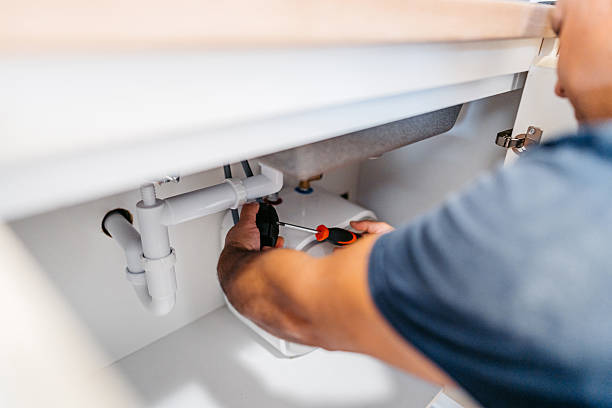 Close-up of a plumber using a screwdriver to tighten a fitting on a white sink drain pipe under a cabinet