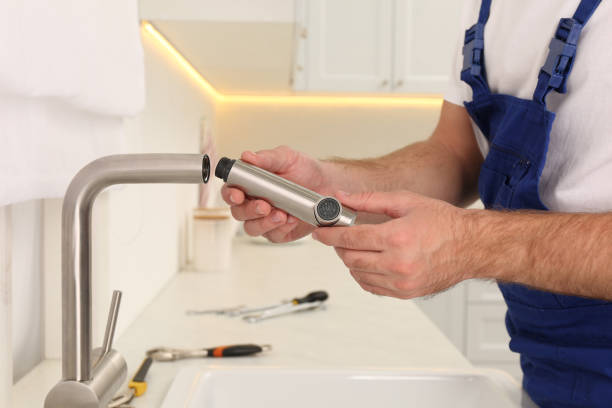 Plumber in blue overalls holding a stainless steel faucet spray head while installing a kitchen faucet