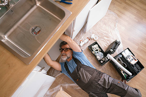 Overhead view of a plumber lying on the floor reaching under a kitchen sink to repair the plumbing, with a toolbox nearby