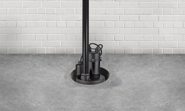 A black submersible sump pump with attached discharge pipe standing on a concrete floor against a brick wall