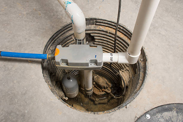 Overhead view of a sump pump basin installed in a concrete floor with pipes, a float switch, and an alarm float visible