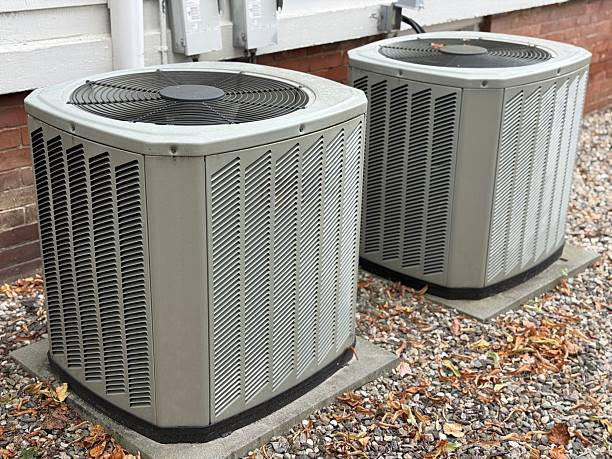 Two outdoor central air conditioner condenser units on gravel beside a brick wall with fallen leaves